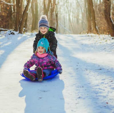 Two children sledding down a snowy path surrounded by trees.